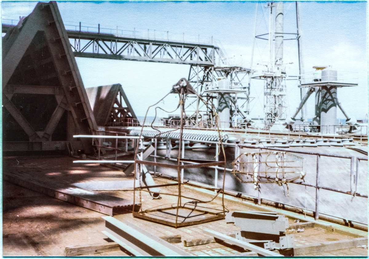 Image 012. Up on the pad deck at Space Shuttle Launch Complex 39-B, Kennedy Space Center, Florida. You are looking northeast, across the Flame Trench with the Flame Deflector topped by its row of Spray Headers filling the view of the Trench, and just left of that, on the far Trench wall, the comb-like row of nozzles on another Spray Header. At far left, shadowed by the RSS and FSS which both loom far above you into the sky just out of frame farther left, one of the large triangular-shaped Side Flame Deflectors sits parked in its Launch Position, and across the Trench in partial sunlight its mate sits in its Park Position. To the right of that, a confused tangle of steel comprises the North Piping Bridge, LH2 Tower, MLP Mount Pedestals, Sound Suppression System Water Tower, SSW supply risers for the MLP, and the Pneumatics Tower, not in that order, but before this is over, you will know them all, well. Center of frame, resting on the pad deck among pieces of loose steel and equipment, the Skip Box which you would ride, lifted by the crane to otherwise inaccessible locations on the tower, sits at the ready. To its right, secured to the removable handrail that guards the five story vertical drop to the bottom of the Flame Trench just beyond it, a stretcher can be seen, also at the ready, in case someone needed evacuation following serious injury. Photo by James MacLaren.